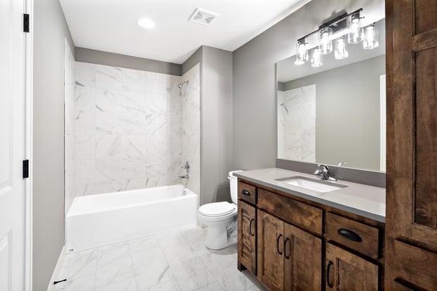 Guest bathroom in a Dusty Rhodes custom home in the Kansas City area, featuring a marble tile tub surround, stained wood vanity, and a glass cylinder light fixture.